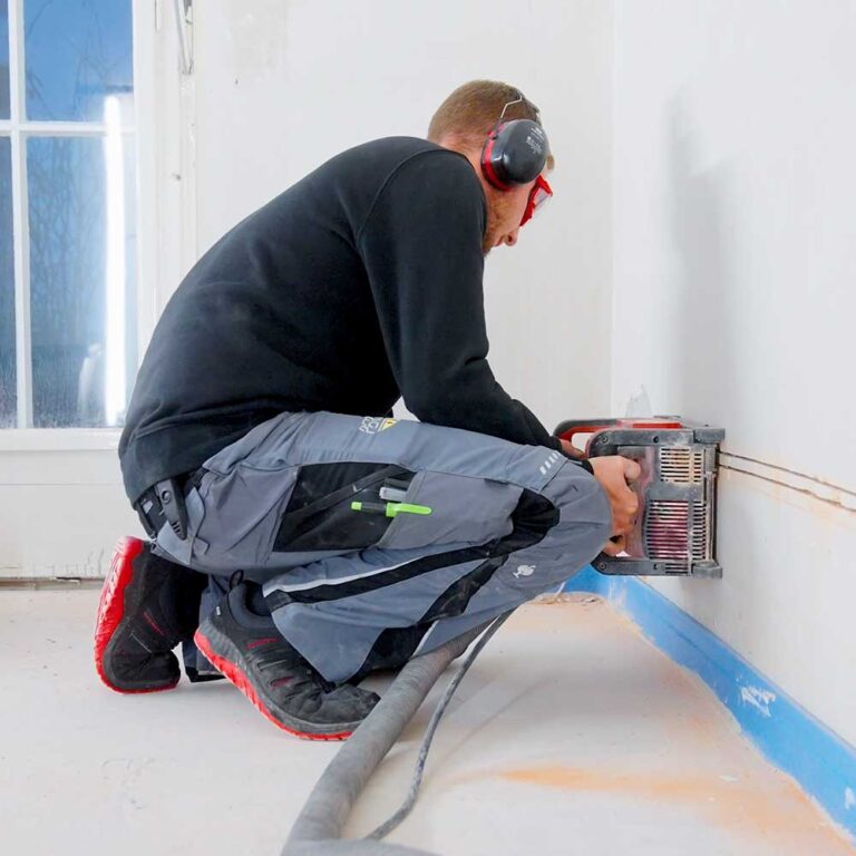 Ein Elektriker bei der Arbeit, der mit einem Schlitzfräse-Werkzeug Kabelkanäle in die Wand schneidet, um die Elektroinstallation in einem Altbau vorzubereiten. Der Techniker trägt Gehörschutz und Schutzbrille, um Sicherheit bei der Arbeit zu gewährleisten.