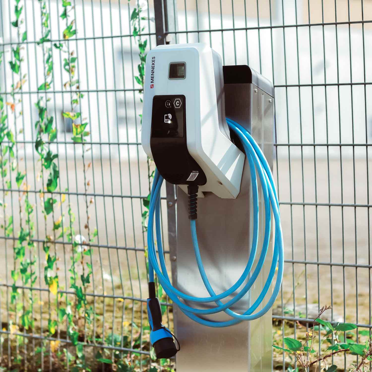 Ladestation für Elektrofahrzeuge der Marke Mennekes, installiert an einem Metallpfosten im Freien, mit aufgewickeltem Ladekabel – Beispiel für moderne Elektroinstallationen zur Integration von E-Mobilität.